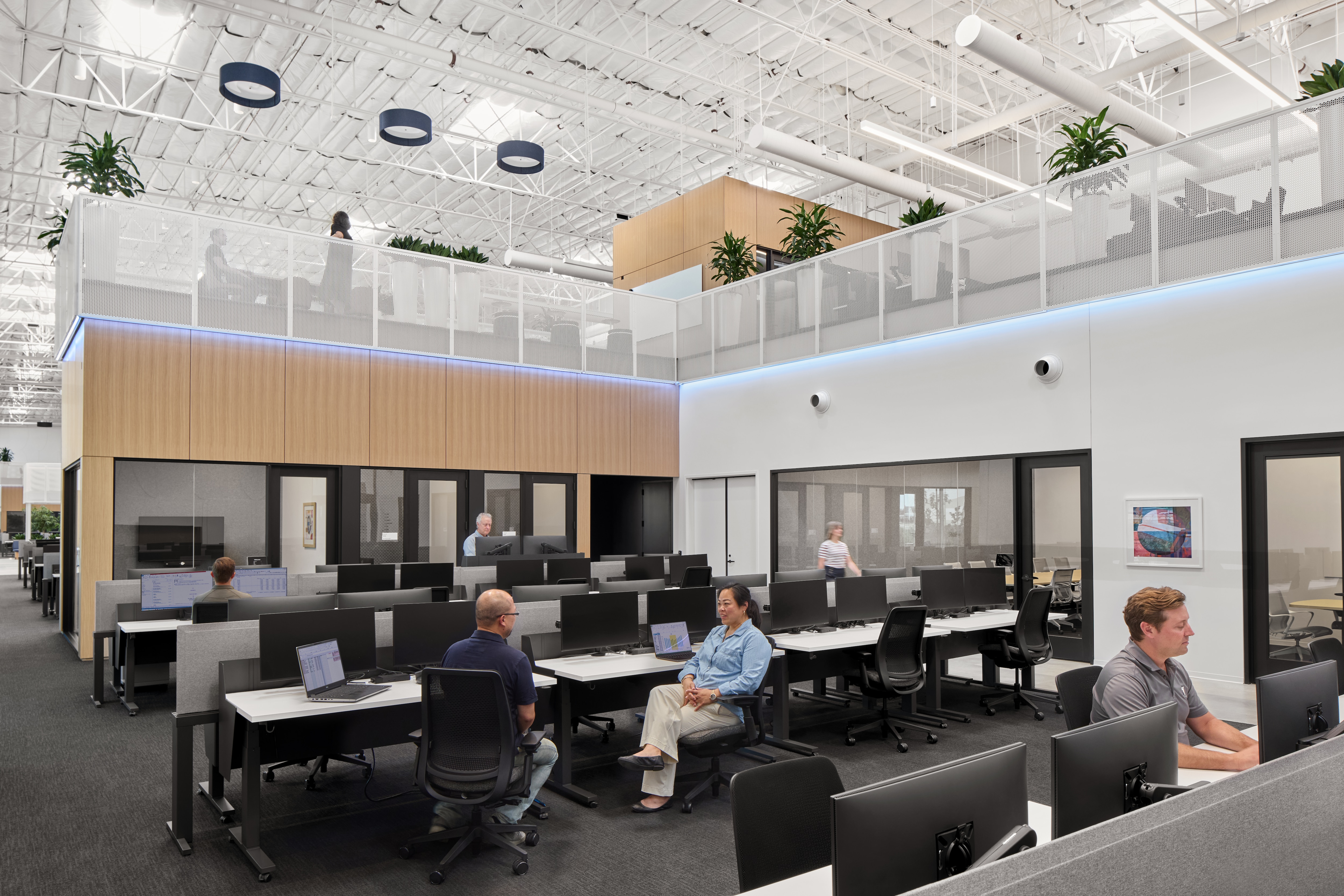 An architectural rendering of an office space with natural wood tones, white walls and ceiling, open space, plant accents and a lot of natural light. People sit along several long desks in a large open area.